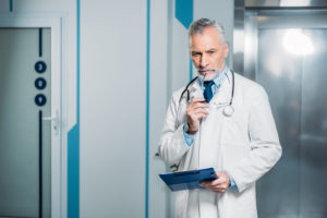 Pensive Mature Male Doctor Holding Eyeglasses And Clipboard In Hospital - Verçosa Contabilidade Inteligência Fiscal LTDA